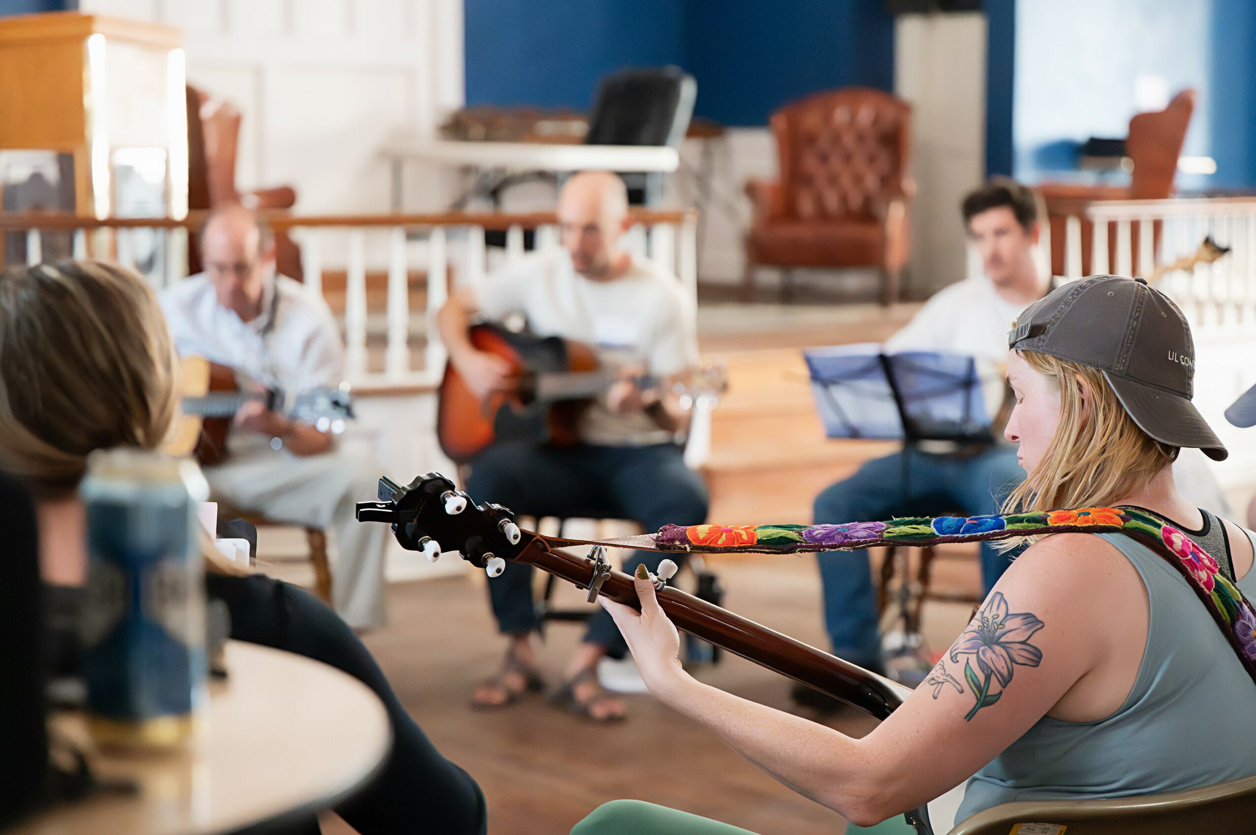 Group of people in a cozy room, some playing guitar and others singing or listening, with a woman in the foreground playing guitar.
