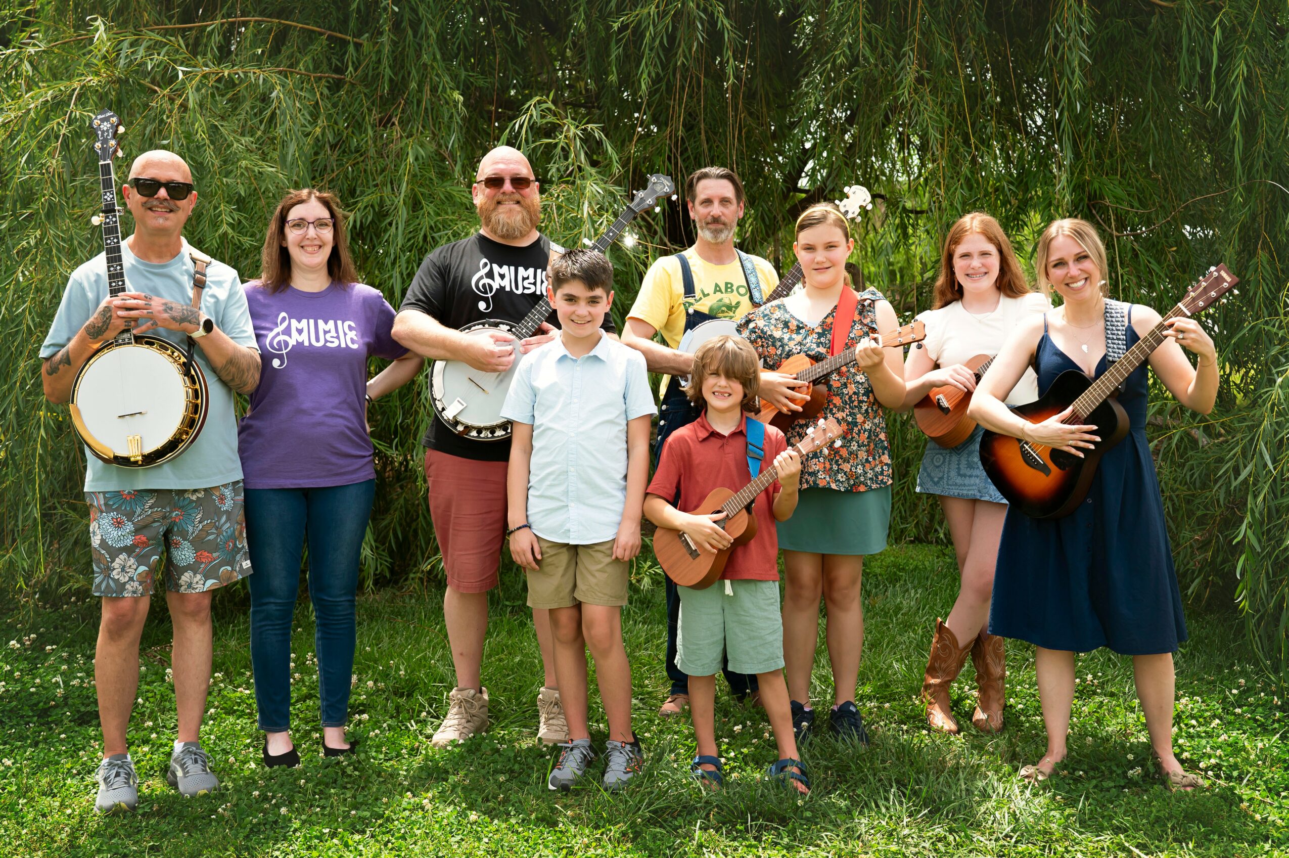 Group of nine people outdoors, some holding musical instruments, standing on grass with trees in background.