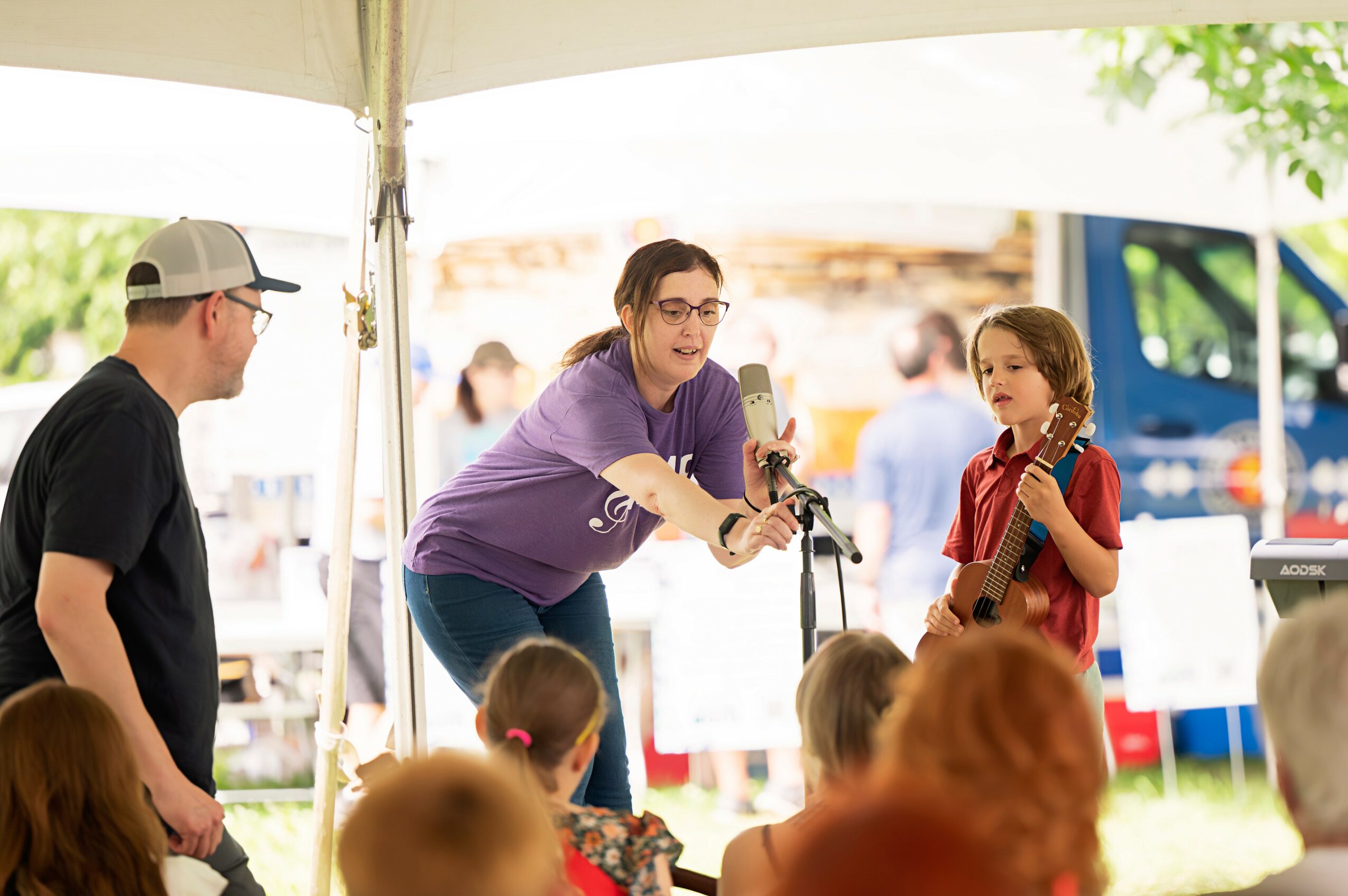 Woman singing into microphone outdoors with children and audience under a tent.
