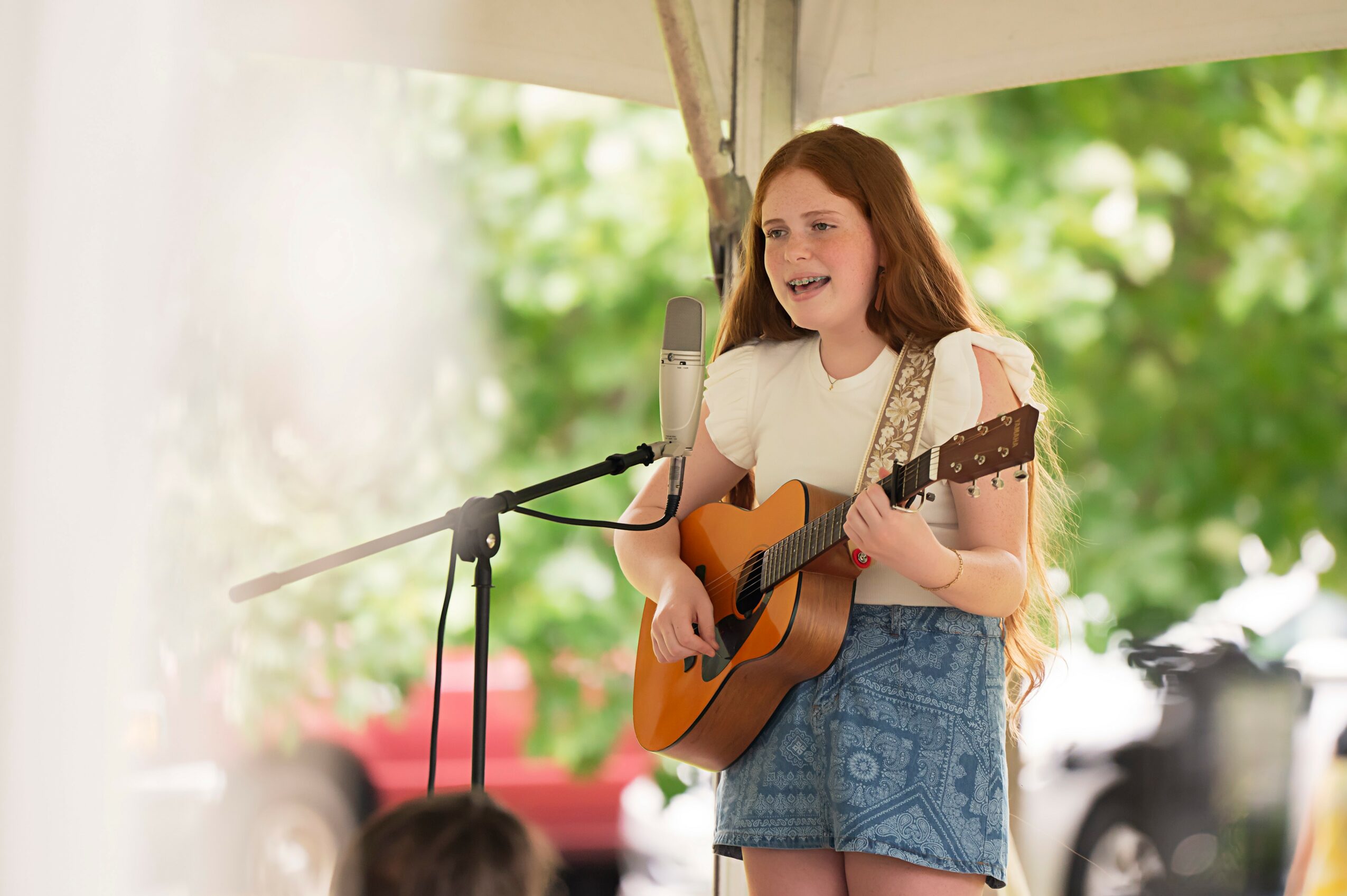 Young girl singing and playing guitar outdoors with a microphone, smiling, surrounded by green trees and cars.