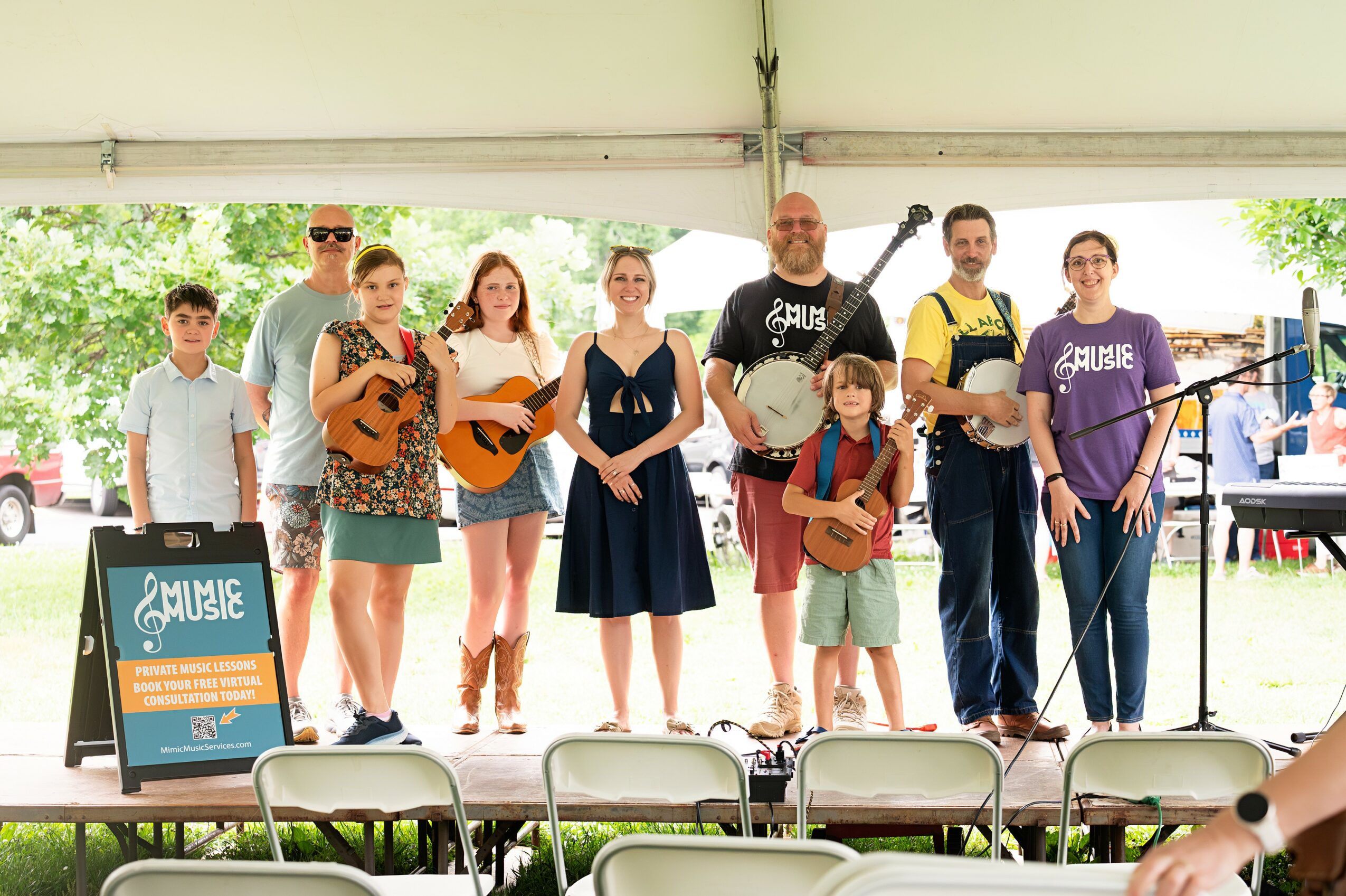 Group of people performing music on stage under a tent, with audience chairs in front, outdoors with trees in background.