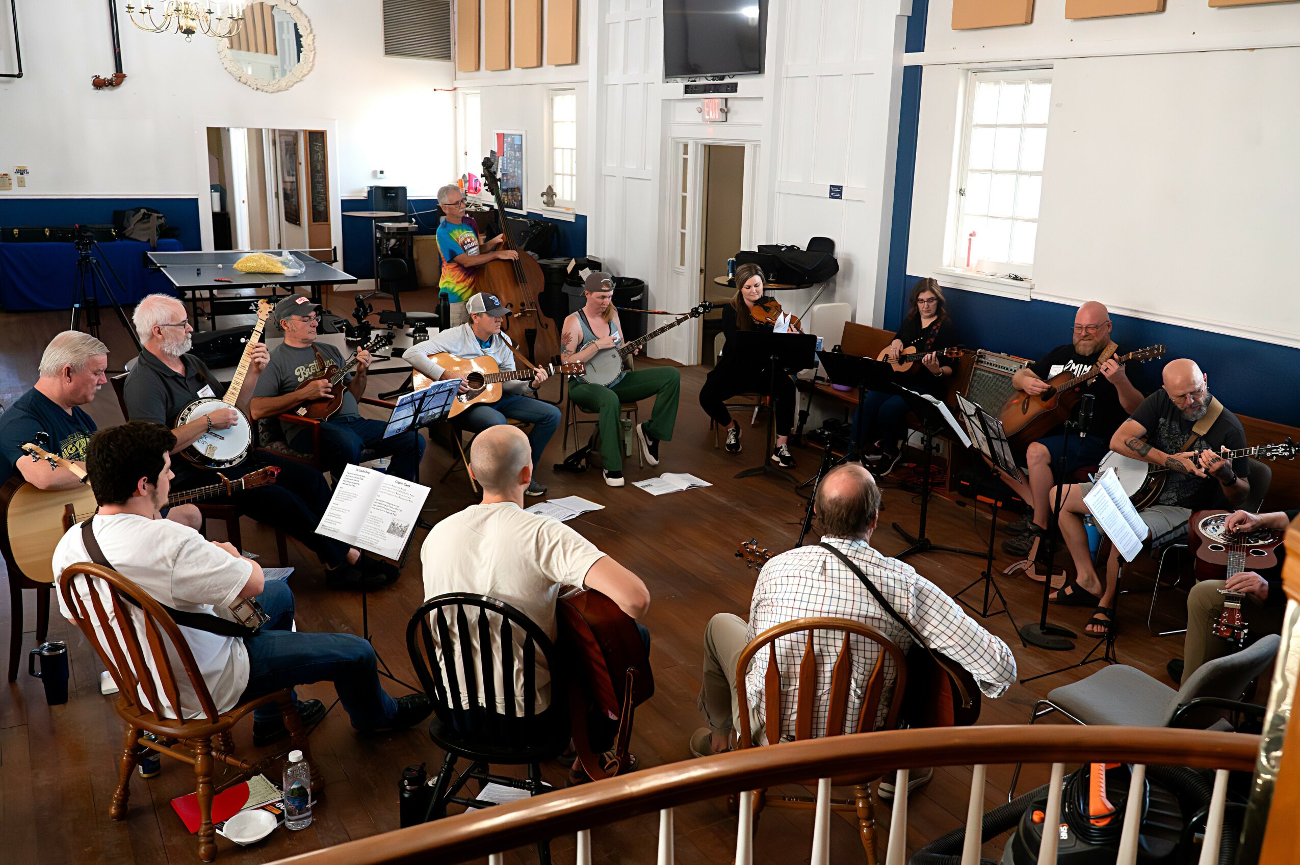 Group of people playing guitars in a room with white walls, windows, and wooden floors, some seated and some standing.