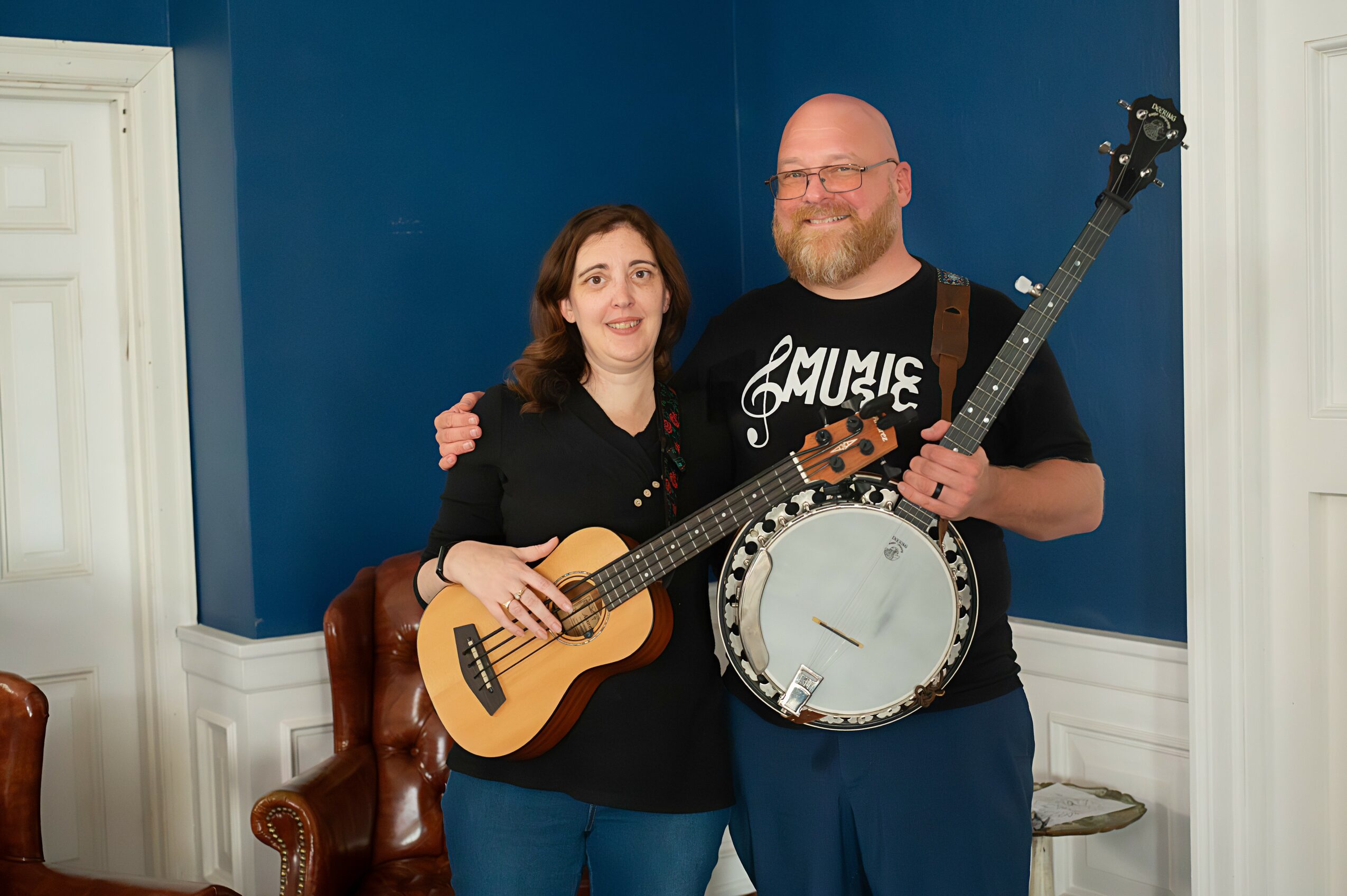 A man and woman stand together indoors, holding musical instruments, smiling at the camera. The man holds a banjo and a guitar.