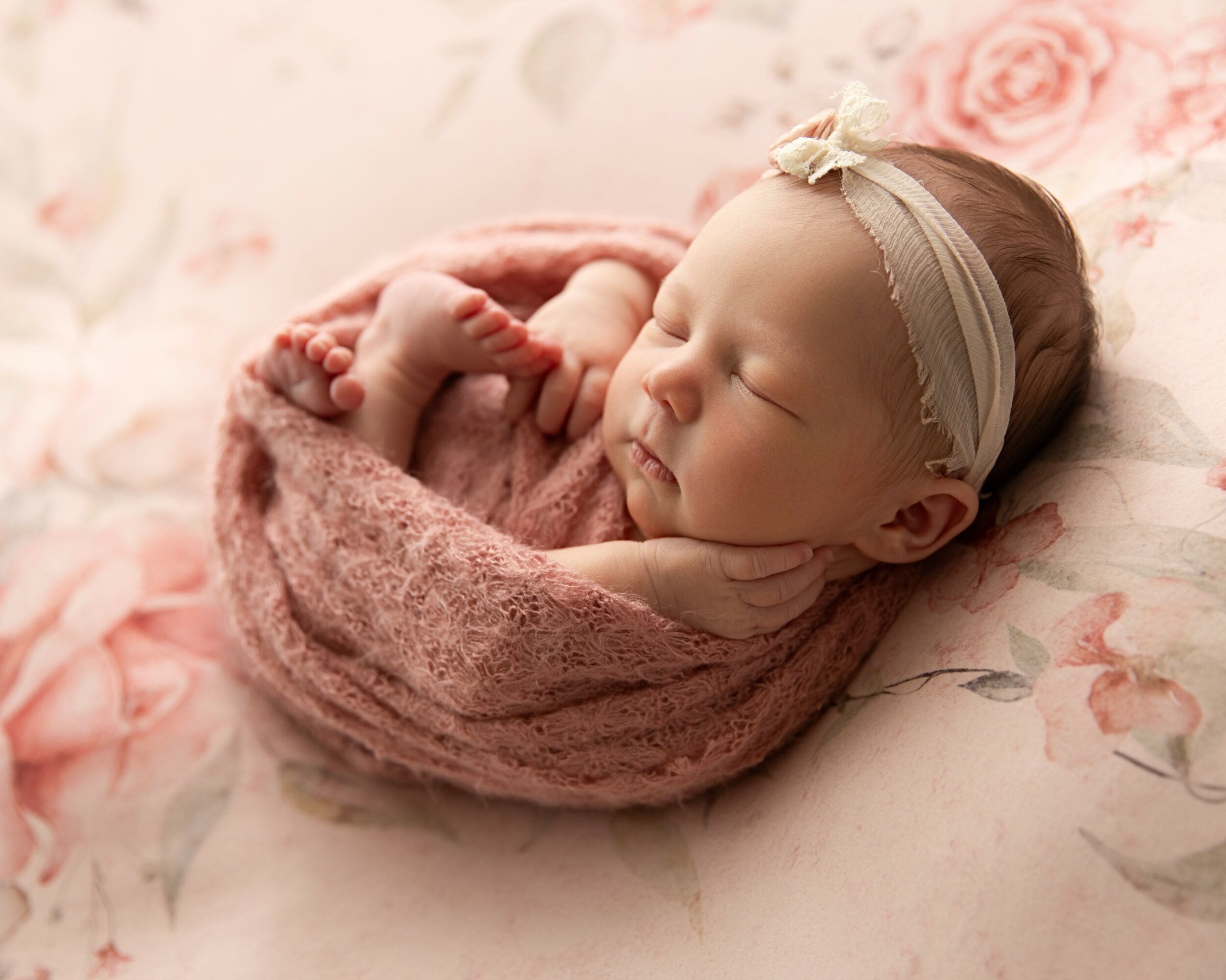 Sleeping newborn baby wrapped in a pink blanket, wearing a headband with a bow, on floral bedding.