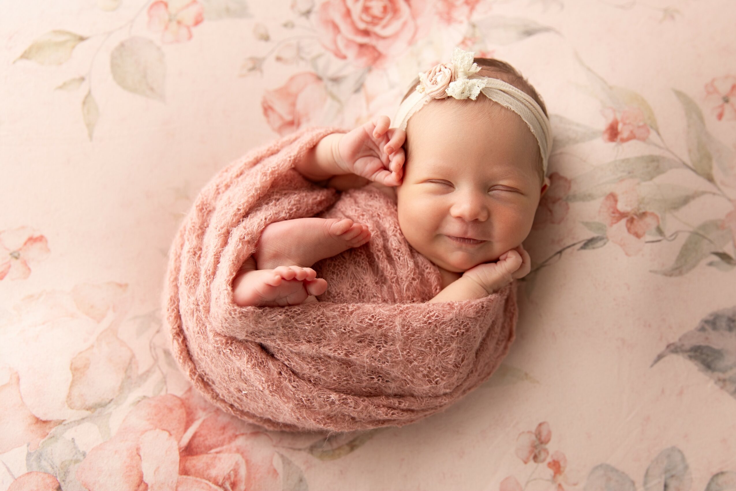 Smiling baby wrapped in pink blanket, lying on floral background, wearing a headband with a bow.