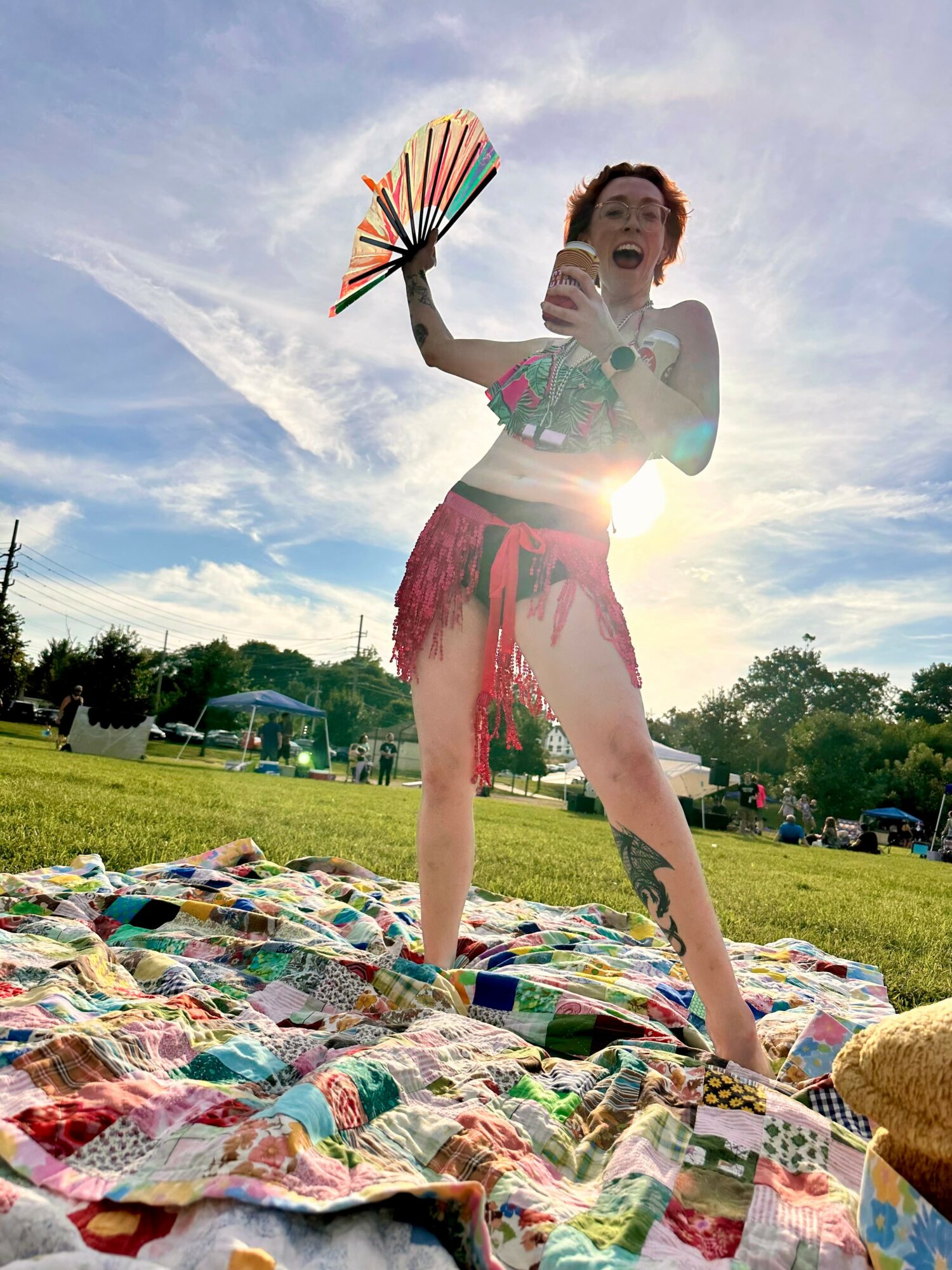 Person standing outdoors on a colorful quilt, holding a fan and a drink, smiling with a bright sky and trees in background.