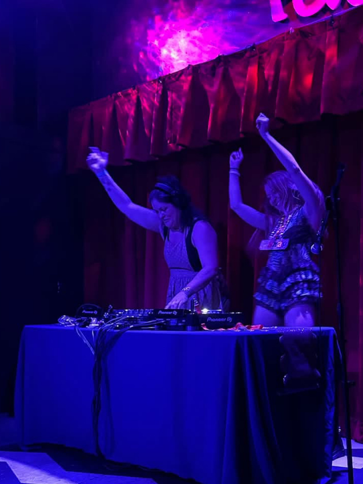 Two women DJing on stage with red curtains and purple lighting, one raising her arm, at a nightclub or event.