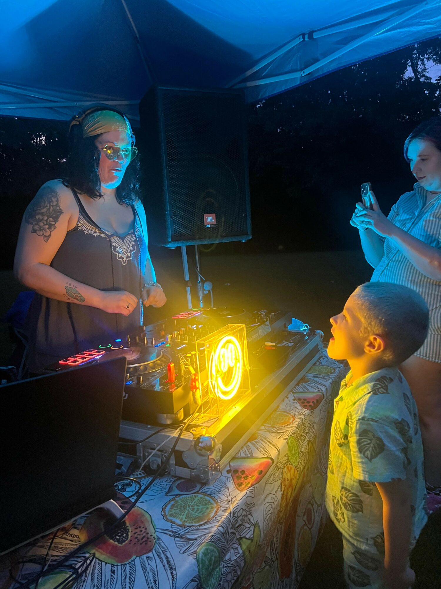 Two people, one with a tattoo, are near a glowing yellow object on a table, with a speaker and equipment nearby.