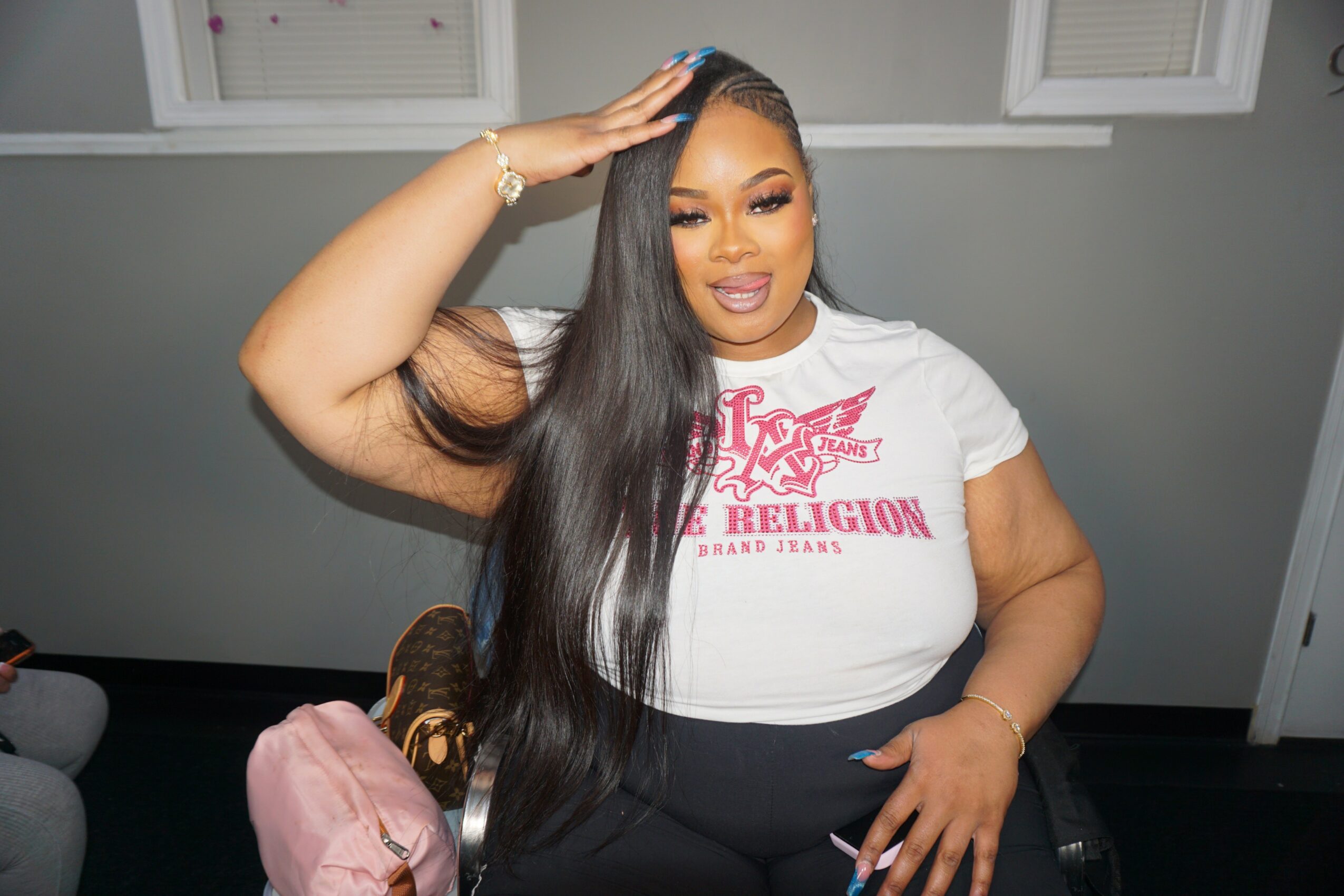 Woman with long black hair, wearing a white T-shirt with pink text, smiling and touching her hair, sitting indoors.