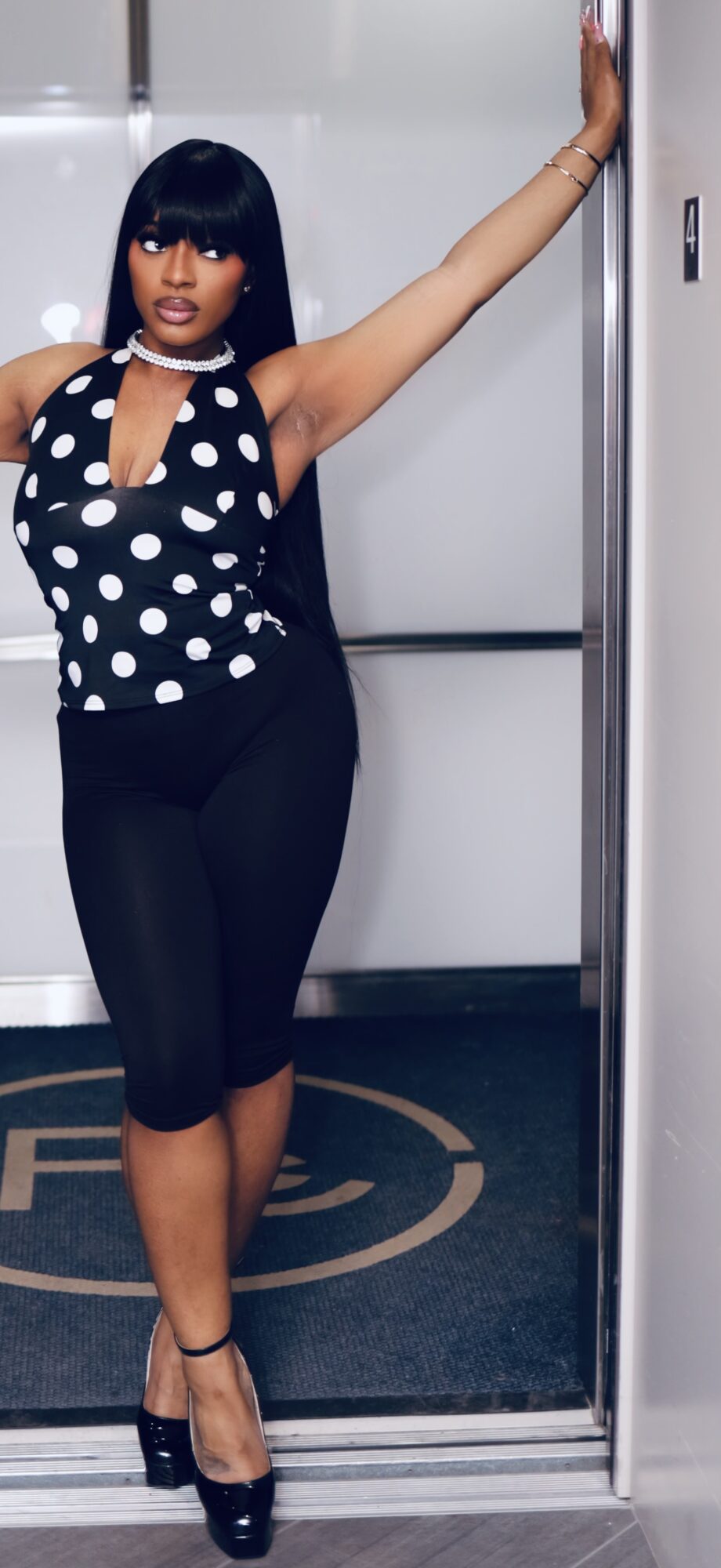 Woman in polka dot top and black pants standing in elevator with arm raised, looking at camera.