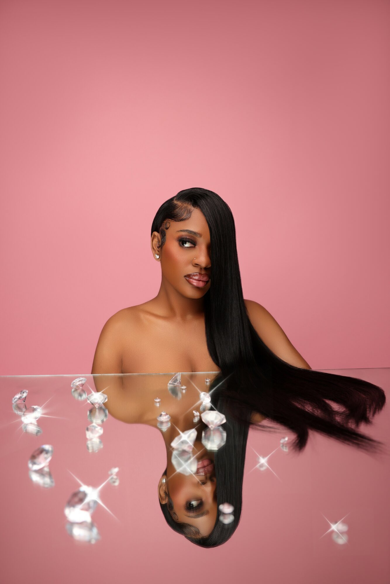 Woman with long black hair and makeup, reflected in water with floating bubbles, against pink background.