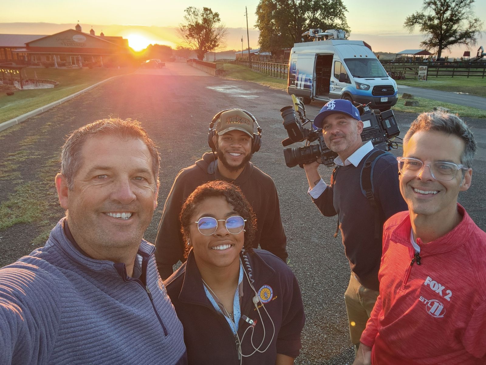 Group of five people outdoors at sunset, smiling, with a van and trees in background.