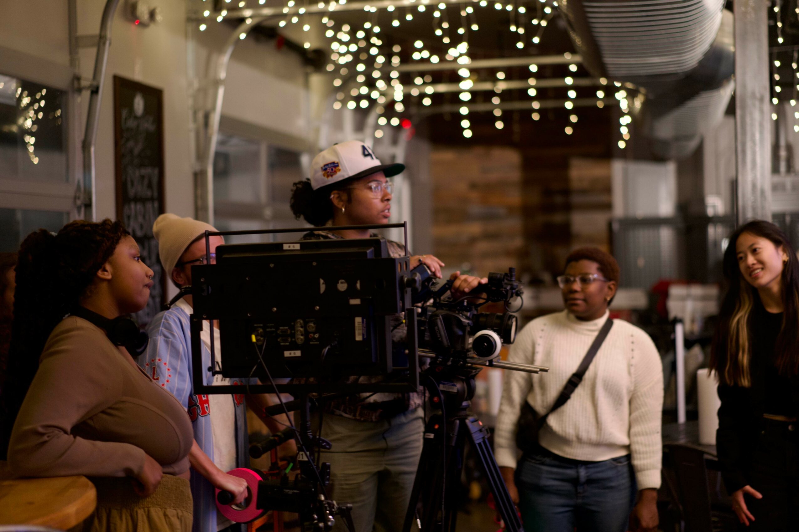 Group of people gathered indoors with string lights, some looking at a camera setup, others talking, in a festive setting.