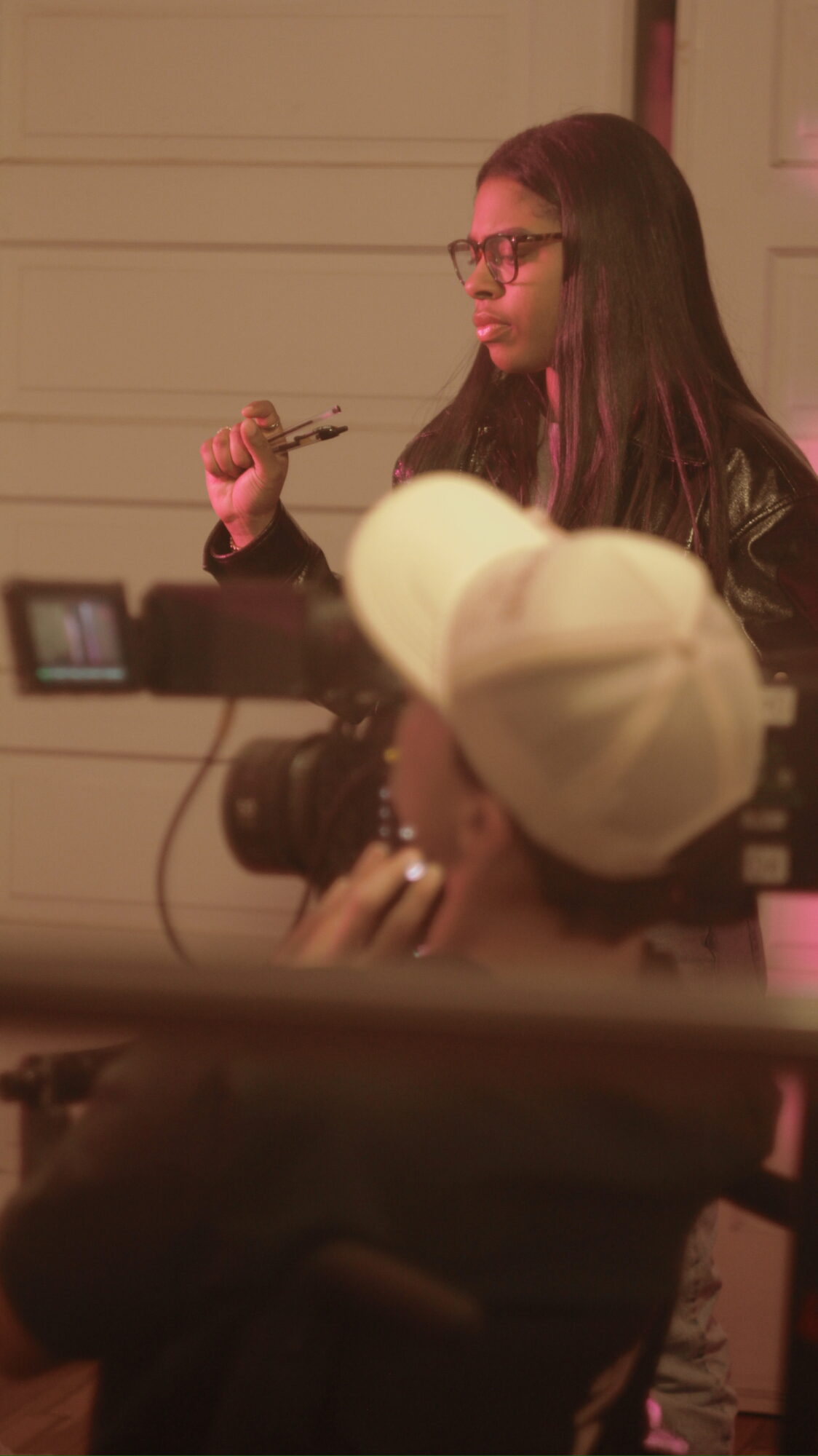 Woman with glasses and long hair holding a pen, sitting in front of a camera, with a person in a white hat nearby.