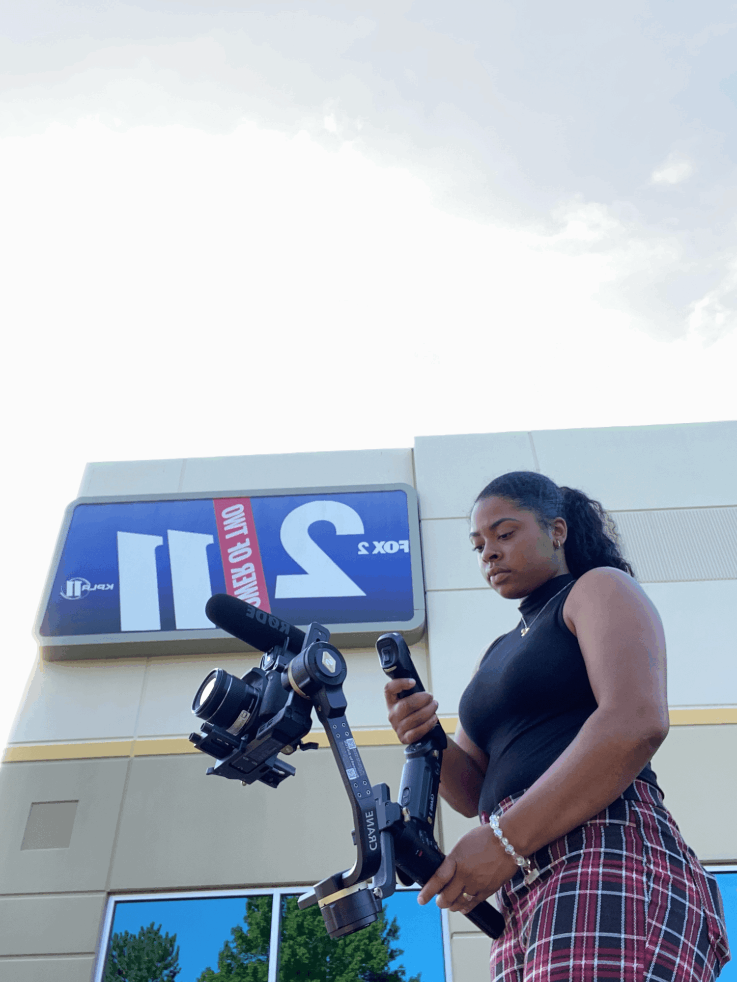 Young woman with a camera and phone standing outside in front of a building with a digital sign, under a cloudy sky.
