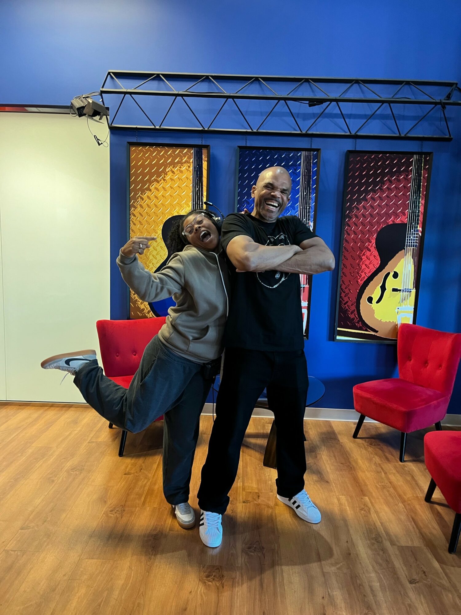 Two people smiling and hugging in a colorful studio with musical instrument decorations, red chairs, and a wooden floor.