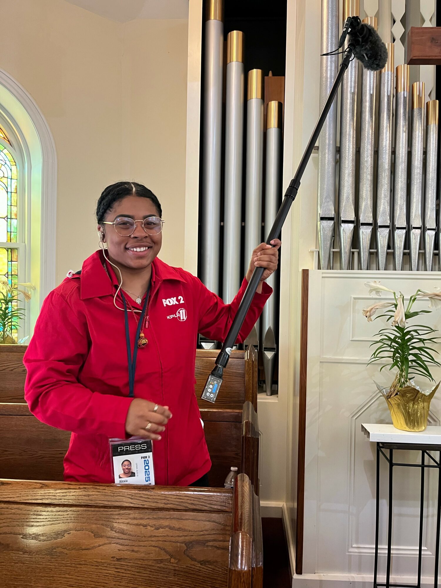 Person smiling, wearing red jacket, holding a long pole with a microphone, standing inside a church near organ pipes.