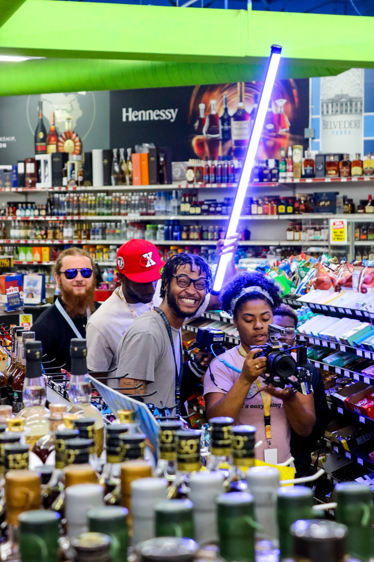 Group of four people shopping in a store aisle with shelves of beverages, one person holding a camera, colorful lighting, and a neon light.