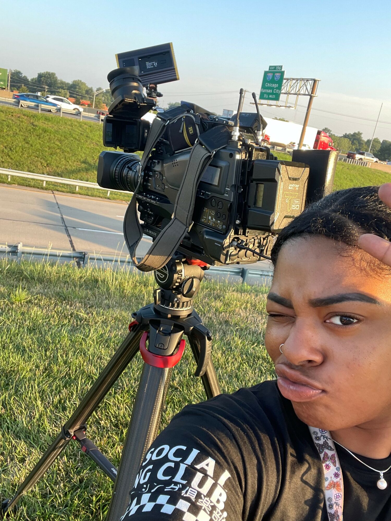 Young person with a camera on a tripod outdoors, winking at the camera, with a highway and green grass in the background.