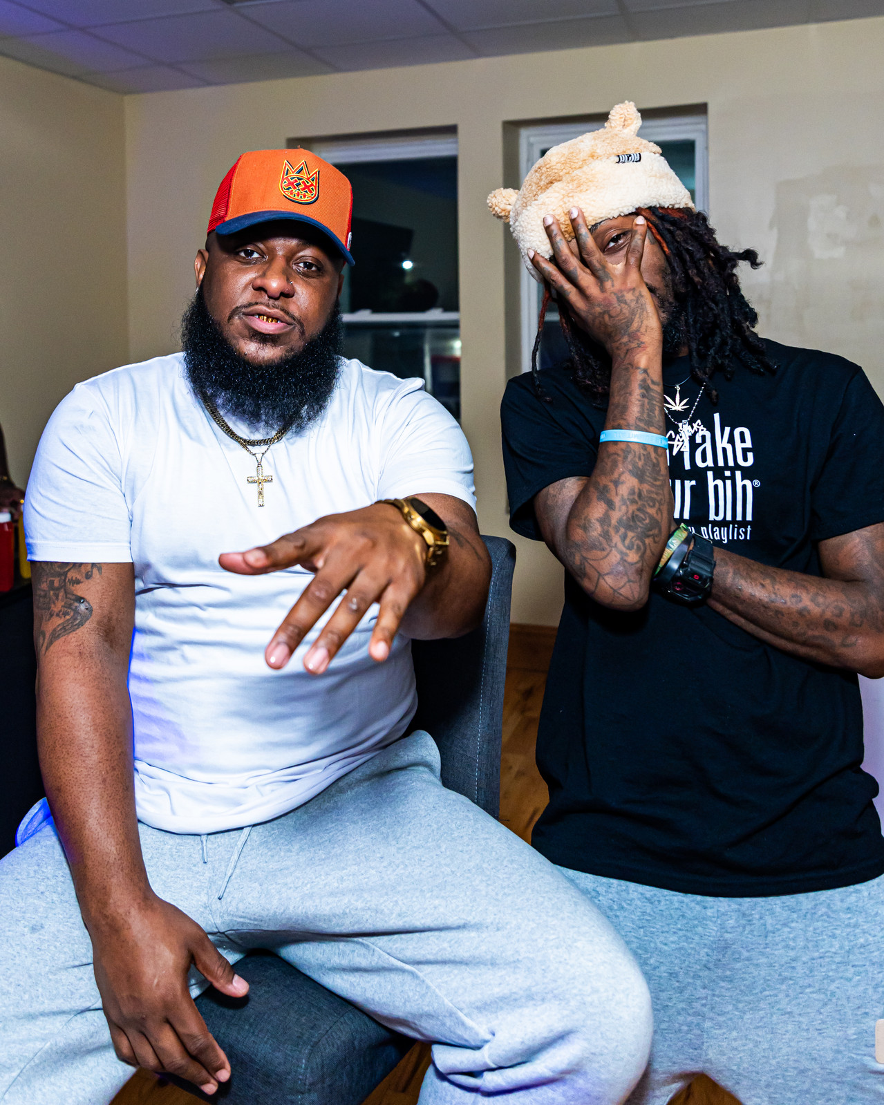 Two men pose indoors, one with a beard and gold chain, the other with dreadlocks and a white hat, covering face.