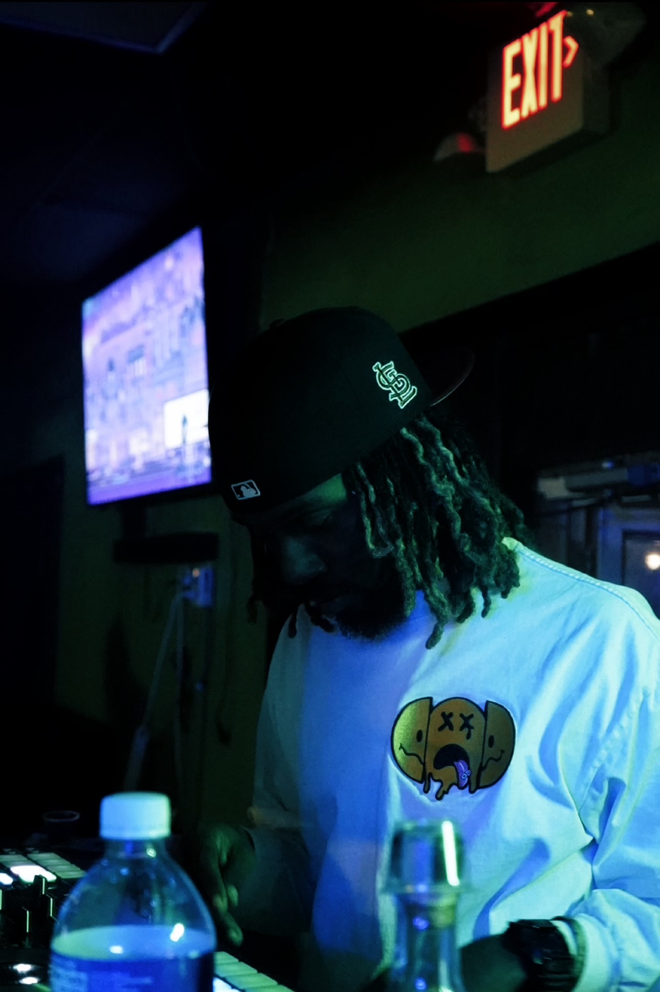 Person with dreadlocks wearing a cap and white shirt in a dimly lit room, with a TV screen and exit sign visible.