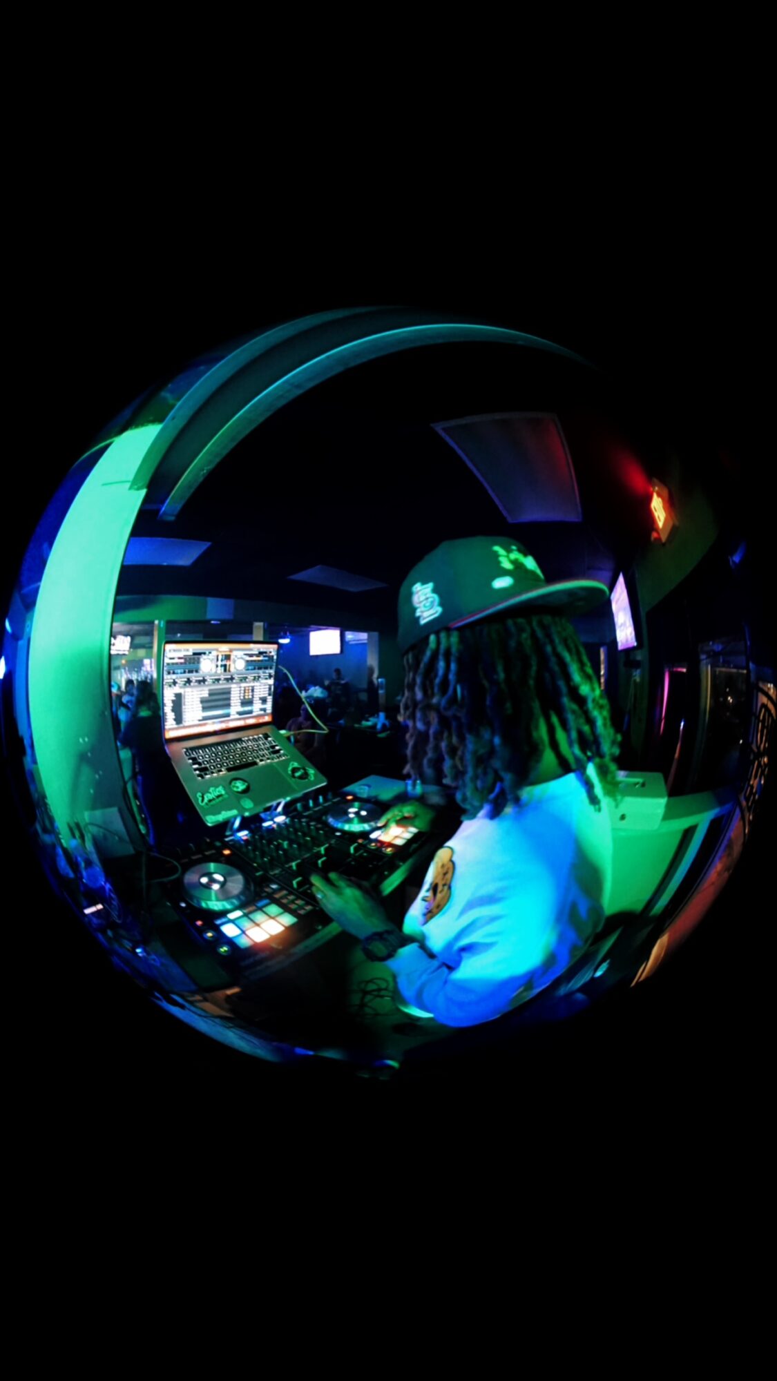 Person with curly hair wearing a hat, working with DJ equipment in a dark room with colorful lights.