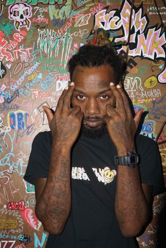 Person with tattoos making a playful face, hands framing face, colorful graffiti background, wearing a black shirt.