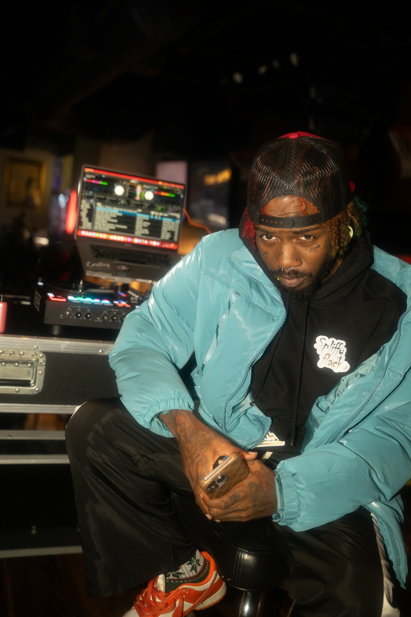 Person with a cap backwards, wearing a blue jacket, sitting in a dimly lit room with DJ equipment and a screen in the background.