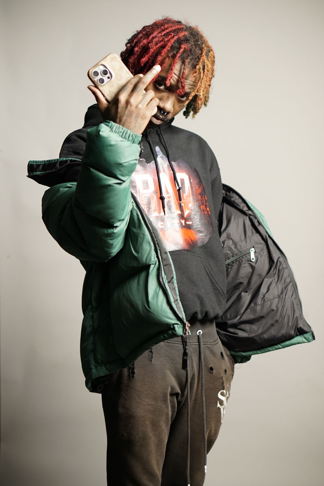 Person with red and blonde hair taking a selfie with a smartphone, wearing a green jacket and black hoodie, against a plain background.