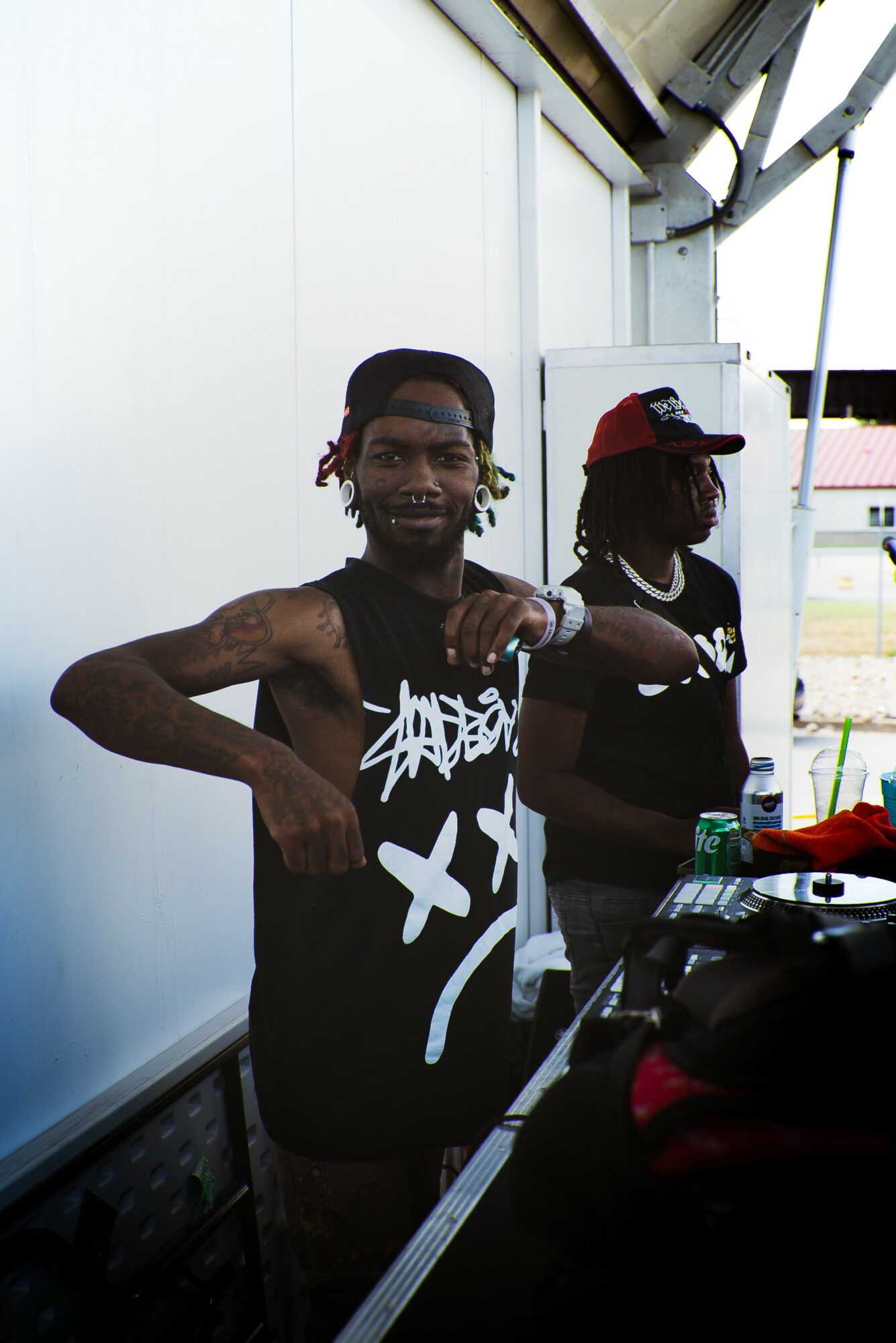 Two young men standing outdoors near a white wall, one wearing a black sleeveless shirt with white graffiti, the other in a red cap and black shirt, with equipment on a table.
