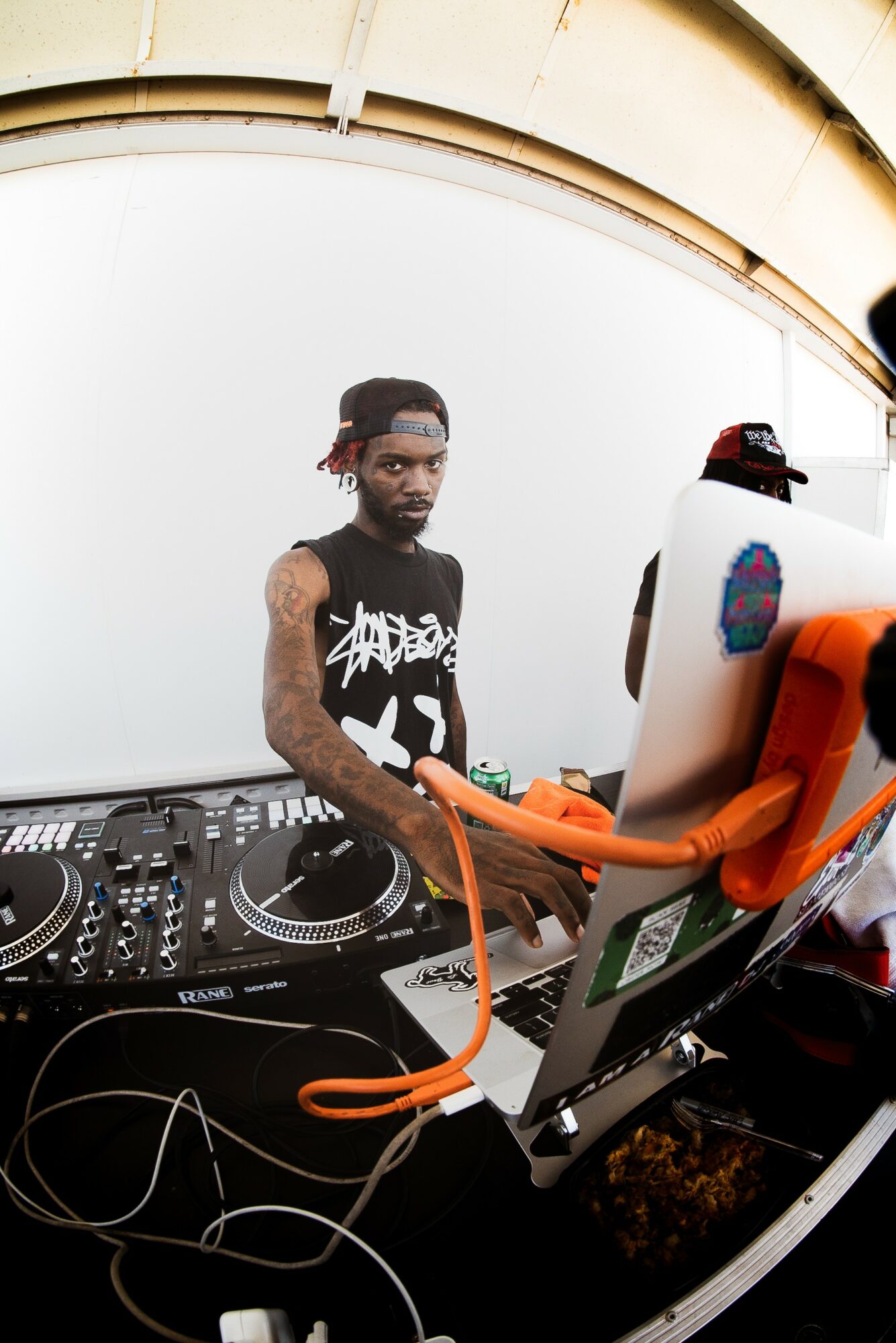 DJ with headphones and equipment, standing under a curved structure, looking at the camera.