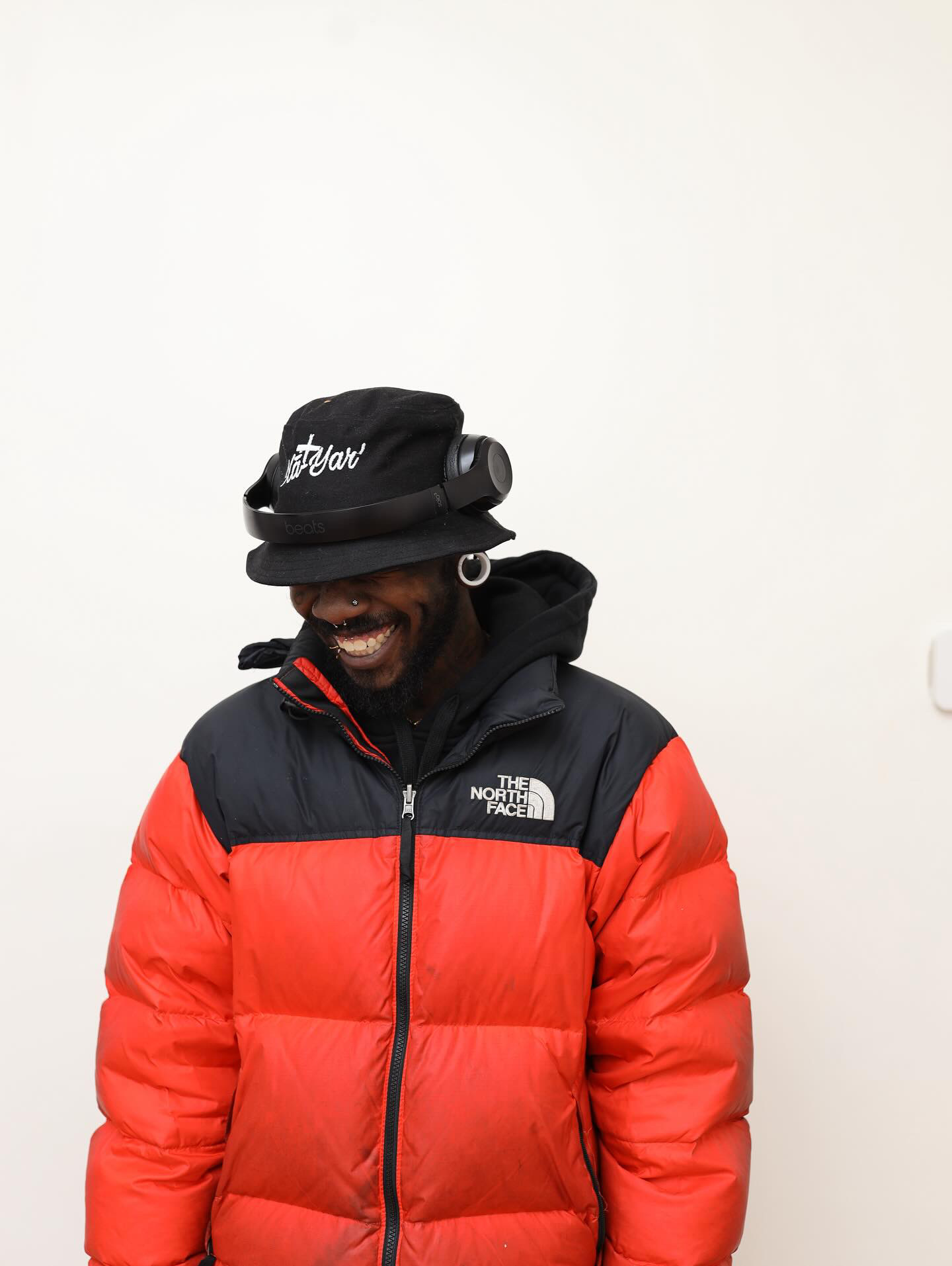 Person wearing a black hat with goggles, red and black jacket, smiling, against a plain white background.