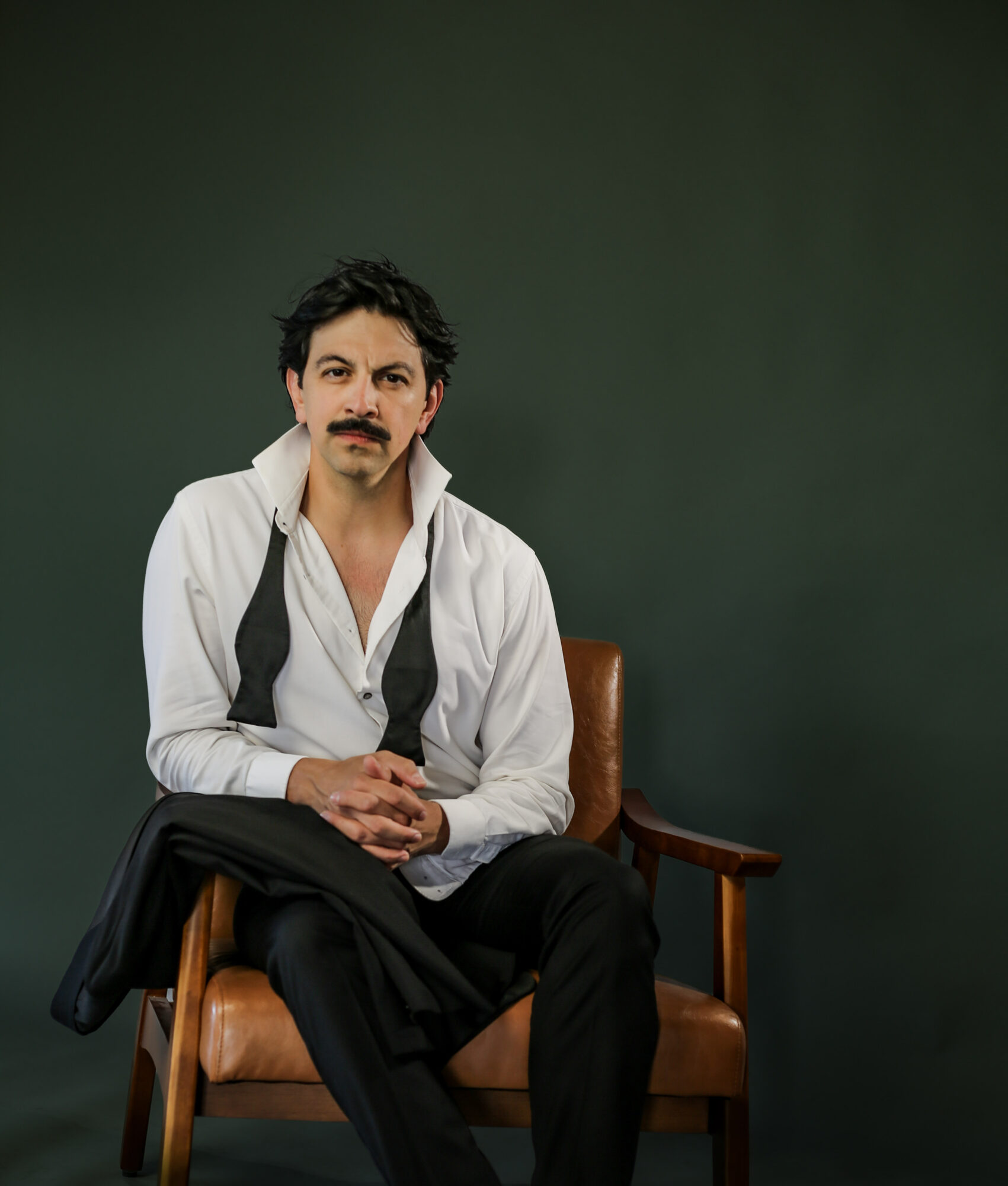 Man with dark hair and mustache sitting on a brown chair against a dark green background, wearing a white shirt and black pants.