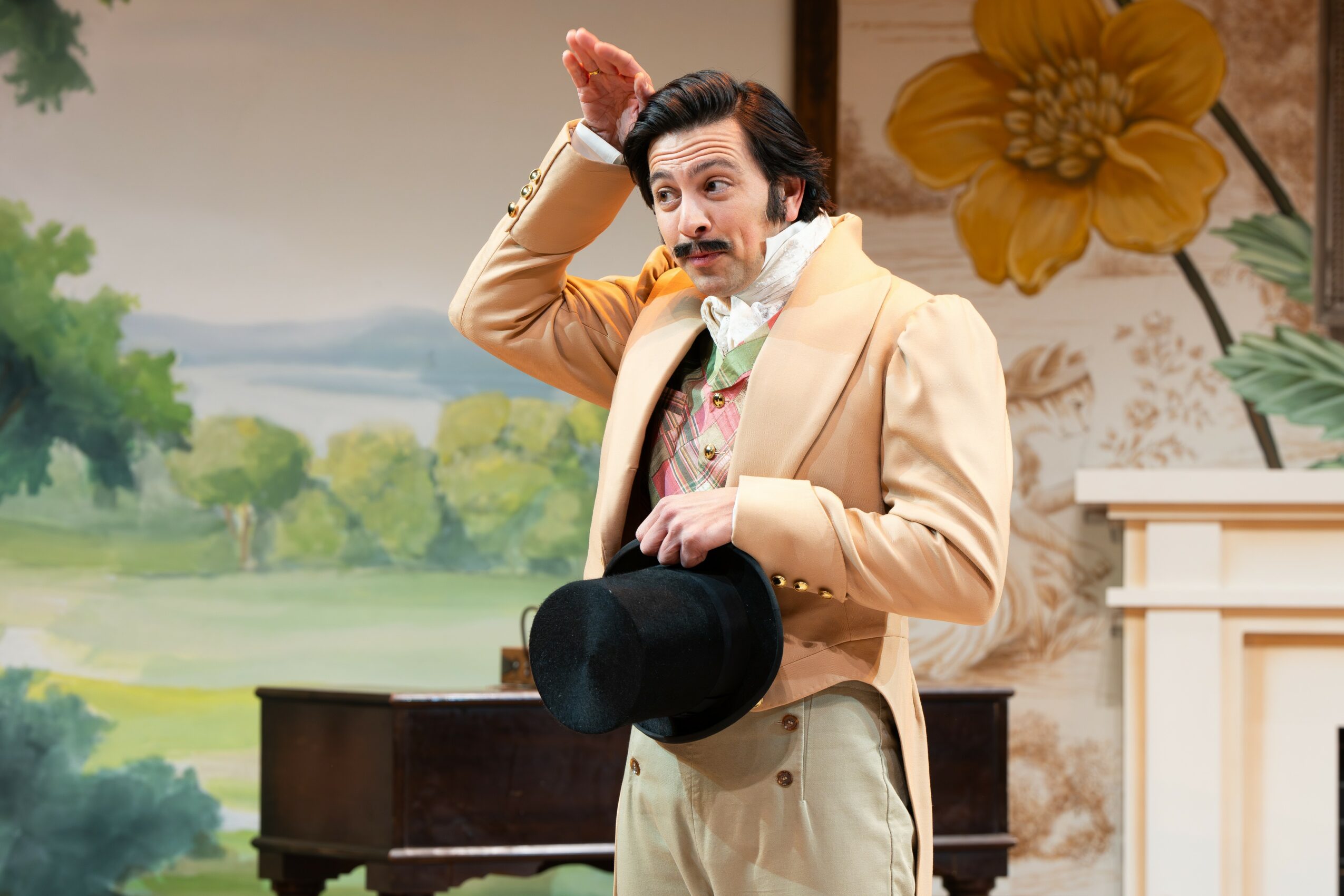 Man dressed in vintage clothing holding a hat, standing in a decorated room with floral and landscape background.